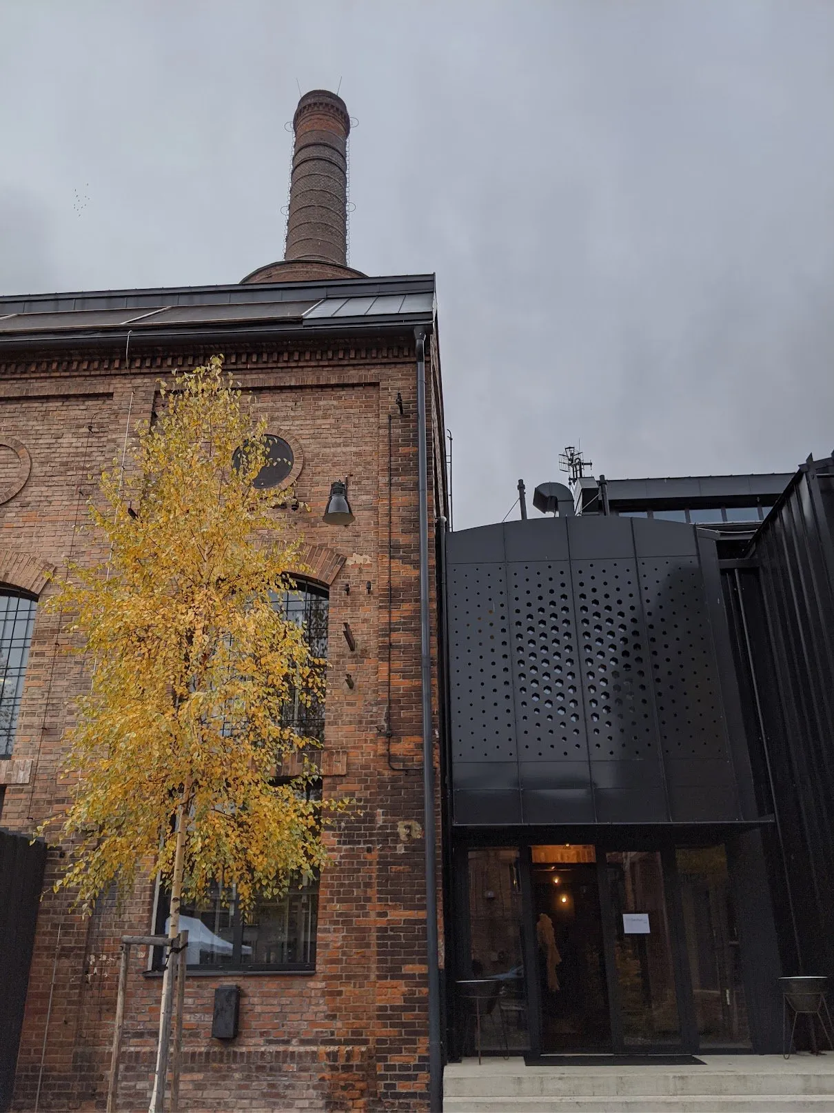 The chimney above a brick hall, with the black perforated-metal entrance building beside it.